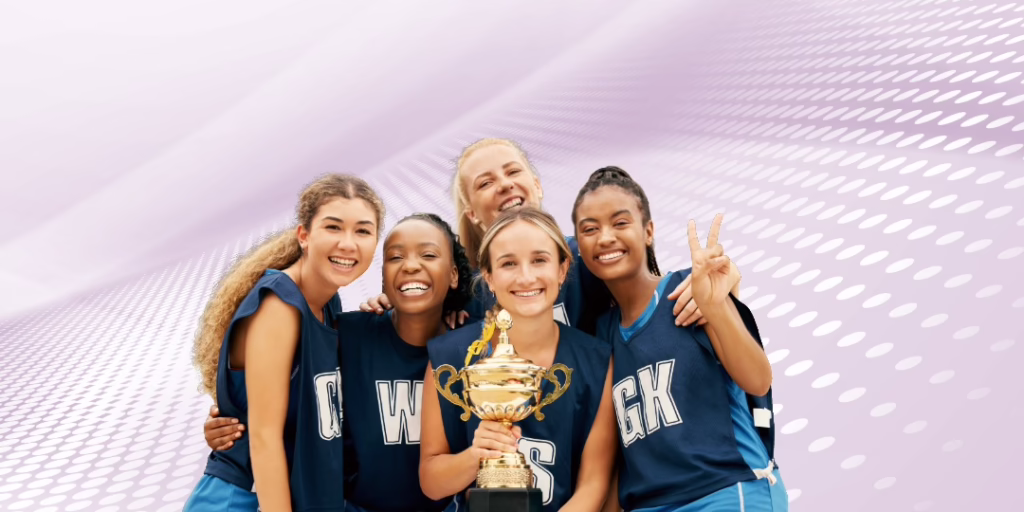 A group of five young women wearing sports jerseys huddle together at a team. The girl in the center holds a shiny gold sports trophy.