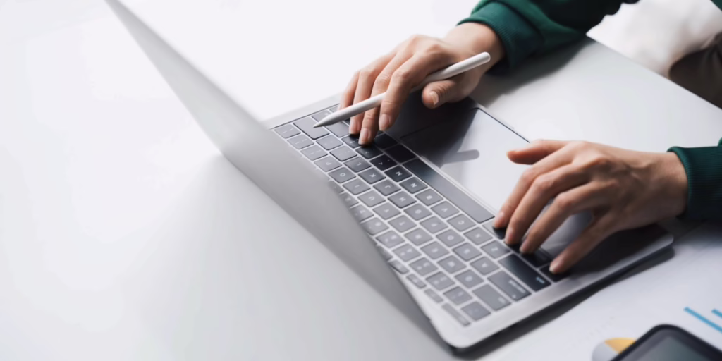 A woman's hands rest on the keyboard of her laptop computer. She is wearing a green shirt and holds a pen in one hand.