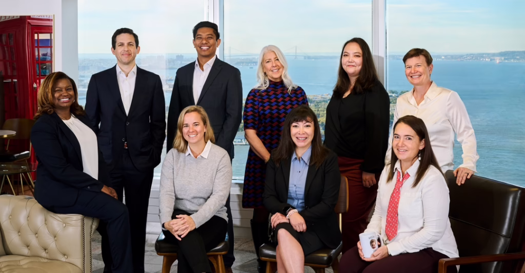 A diverse group of smiling people gathered together in a group. The window behind them looks out at New York Harbor.